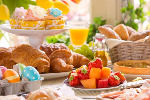 A colorful brunch spread with pastries, cupcakes, decorated eggs, fruit, and juice, set on a table with flowers.