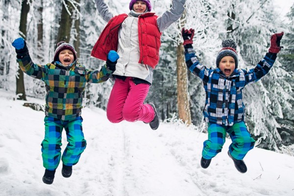Three people in colorful winter clothing are joyfully jumping in a snowy forest, surrounded by snow-covered trees.