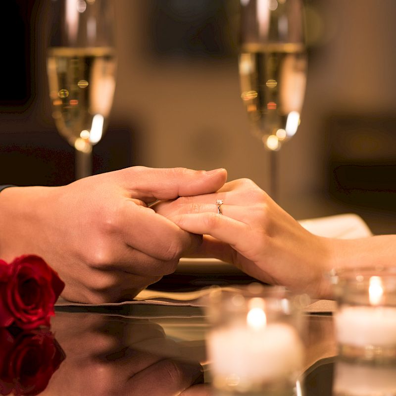 A romantic setting with two people holding hands, a red rose, candles, and two glasses of champagne on a table.