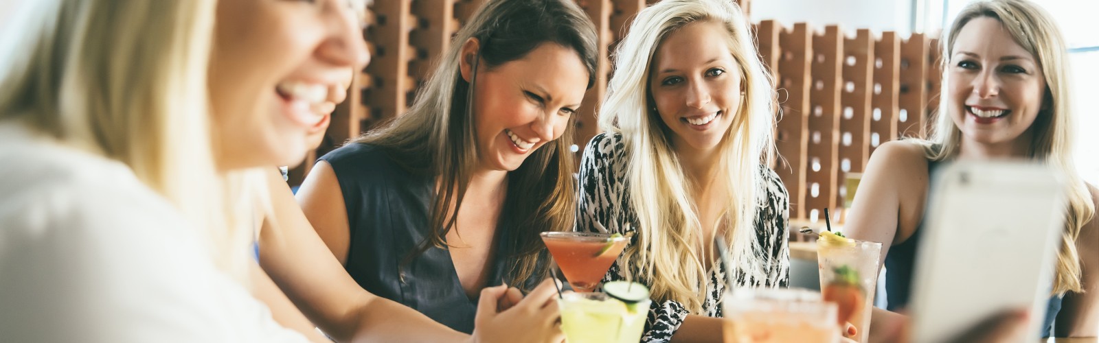 A group of women are sitting at a table, enjoying drinks and laughing while taking a selfie, with a focus on fun and camaraderie.