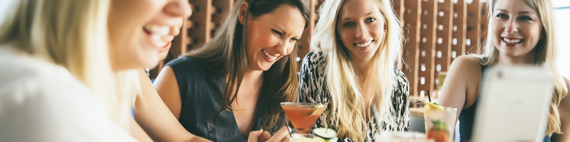 A group of women are sitting at a table, enjoying drinks and laughing while taking a selfie, with a focus on fun and camaraderie.