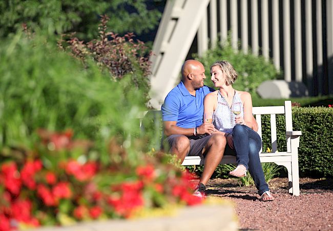 A couple sits on a white bench in a garden, surrounded by colorful flowers and greenery, smiling and holding wine glasses.