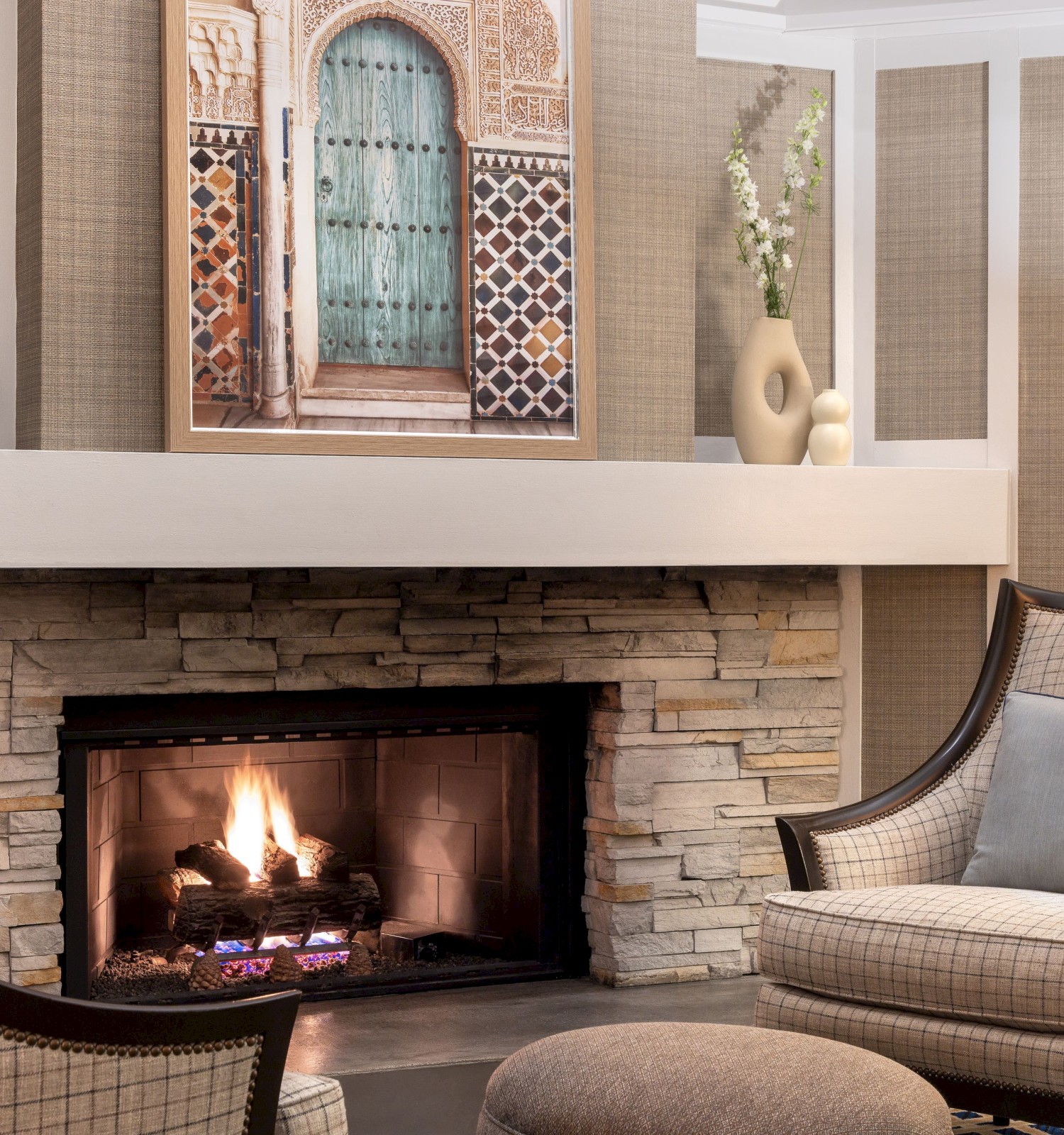 A cozy living room with a lit fireplace, armchairs, a side table, and decorative vases and artwork on the mantel.