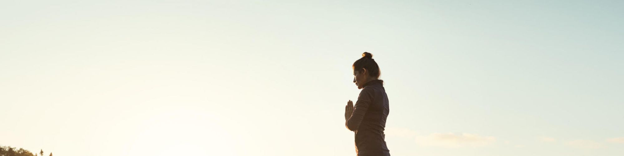 A person stands on a paddleboard on calm water during sunset, holding a yoga pose with hands in a prayer position, in a serene outdoor setting.
