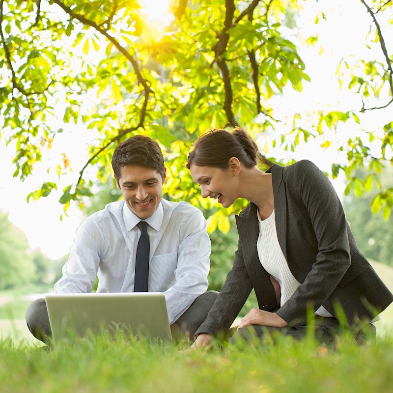 Two people in business attire are sitting on grass under a tree, smiling and looking at a laptop. The setting is outdoors in a park, ending the sentence.