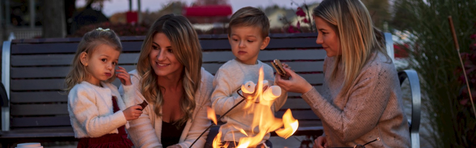 A group of four people, including two children and two adults, sit around a fire pit roasting marshmallows, with Starbucks cups in the foreground.