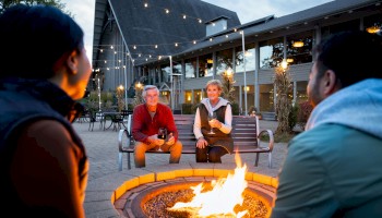 People sitting around a fire pit in an outdoor setting with lights strung above and a building in the background.