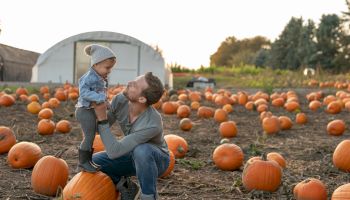 A man kneels, holding a child in a pumpkin field. They appear to be enjoying a moment together surrounded by many pumpkins.