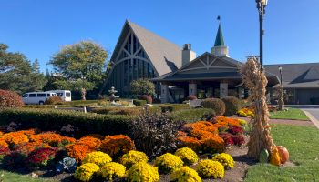 A building with a triangular roof and a landscaped garden featuring vibrant flowers and a lamppost decorated with corn stalks and pumpkins is visible.