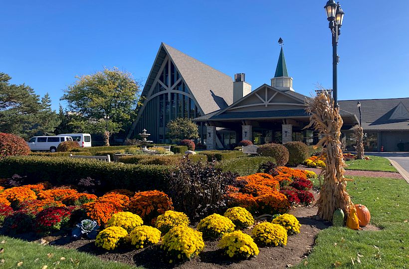 A building with a triangular roof and a landscaped garden featuring vibrant flowers and a lamppost decorated with corn stalks and pumpkins is visible.