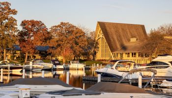 The image shows a marina with docked boats and a large A-frame building in the background, surrounded by trees with autumn foliage.