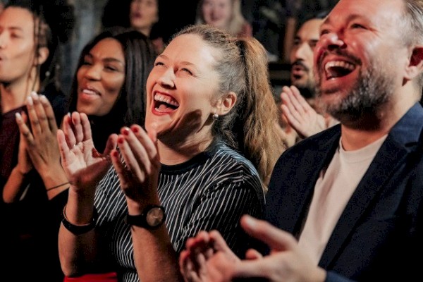 A diverse group of people is applauding and laughing, seemingly enjoying a performance or event. They appear to be in a lively, engaging atmosphere.