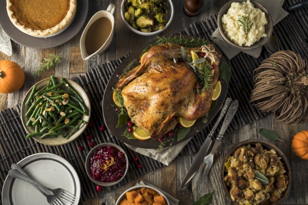 A table is set with a roasted turkey, mashed potatoes, stuffing, green beans, cranberry sauce, gravy, sweet potatoes, Brussels sprouts, and pumpkin pie.
