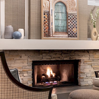A cozy living room with a lit stone fireplace, two chairs, a round ottoman, decorative vases, and a vibrant picture above the mantel.