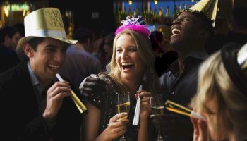 A group of people is celebrating New Year's Eve, wearing party hats, blowing noisemakers, and holding glasses of champagne.