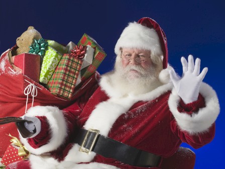 A person dressed as Santa Claus is holding a sack full of wrapped presents, waving with a gloved hand, against a blue background.