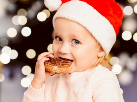 A child wearing a Santa hat is enjoying a pastry with holiday lights blurred in the background.