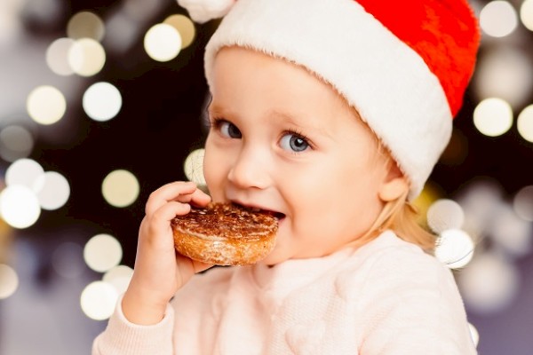 A child wearing a Santa hat is enjoying a pastry with holiday lights blurred in the background.