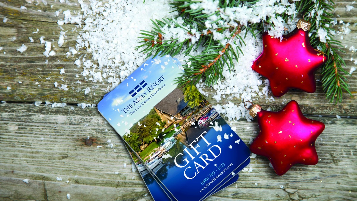 The image shows gift cards, fake snow, pine branches, and two red star ornaments on a wooden surface, creating a festive holiday scene.