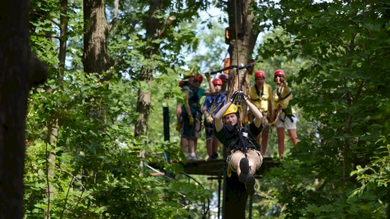 A person is zip-lining through a forest while a group waits on a platform in the background, all wearing helmets and harnesses.