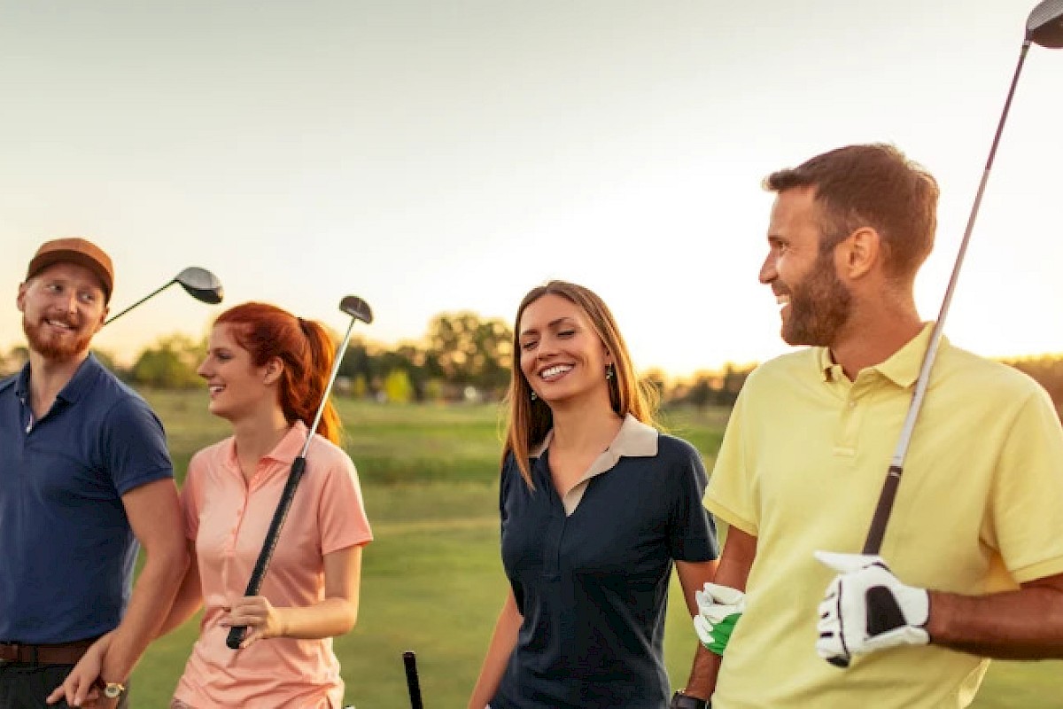 A group of four people smiling and holding golf clubs, walking on a golf course under a warm sunset.