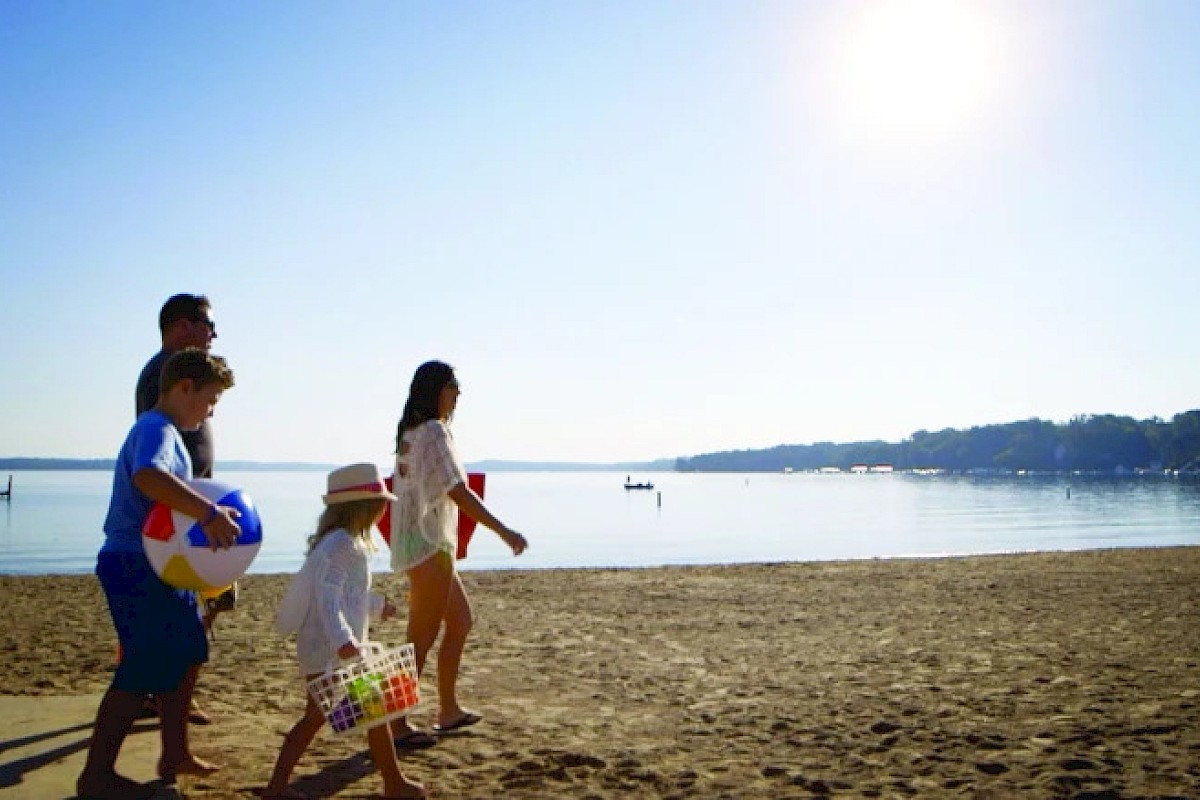 A family walks on a sunny beach, carrying beach balls and a cooler, with a calm sea and clear blue sky in the background.