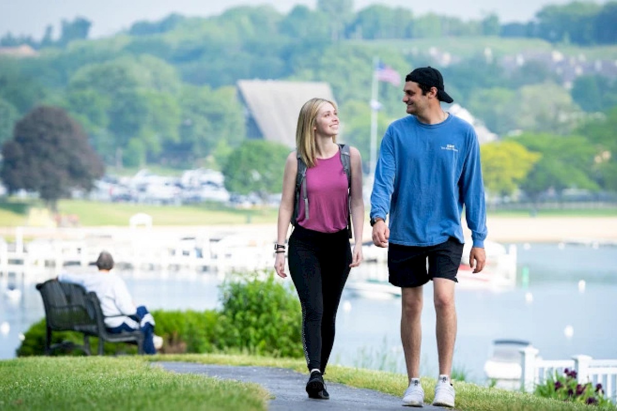 A couple is walking along a scenic lakeside path, surrounded by lush greenery, with a serene view and a person seated on a bench nearby.