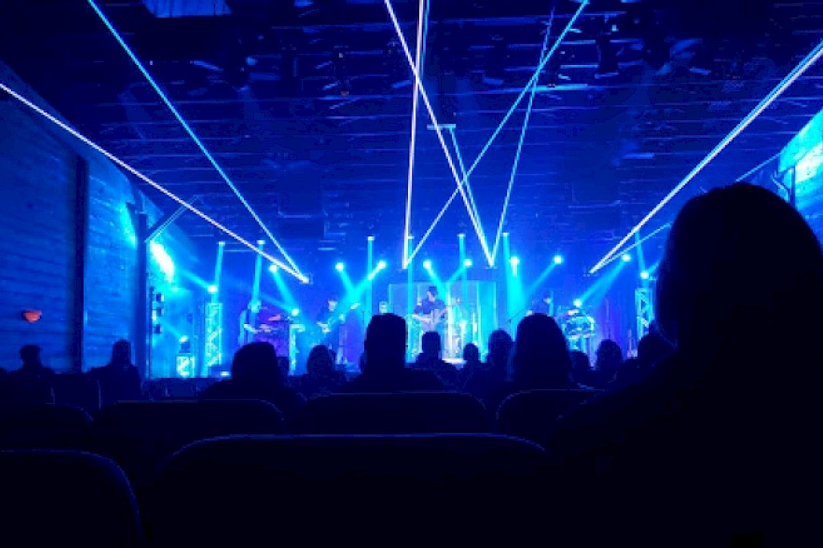 A live concert with a stage featuring blue lighting and laser beams, viewed by an audience seated in a dimly lit venue.