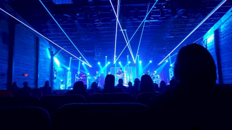 A live concert with a stage featuring blue lighting and laser beams, viewed by an audience seated in a dimly lit venue.