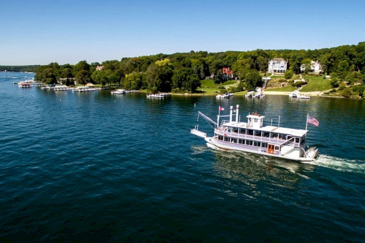 A boat cruises on a lake near a shoreline with trees and houses under a clear blue sky.