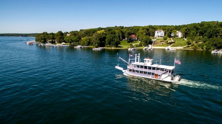 A boat cruises on a lake near a shoreline with trees and houses under a clear blue sky.