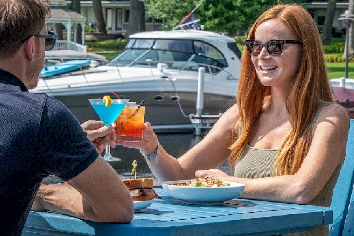 Two people are seated outdoors at a table by the water, enjoying drinks and food, with boats docked in the background.