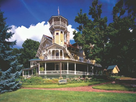 A large, ornate Victorian-style house with a wraparound porch and tower, surrounded by trees and a well-kept lawn under a blue sky.