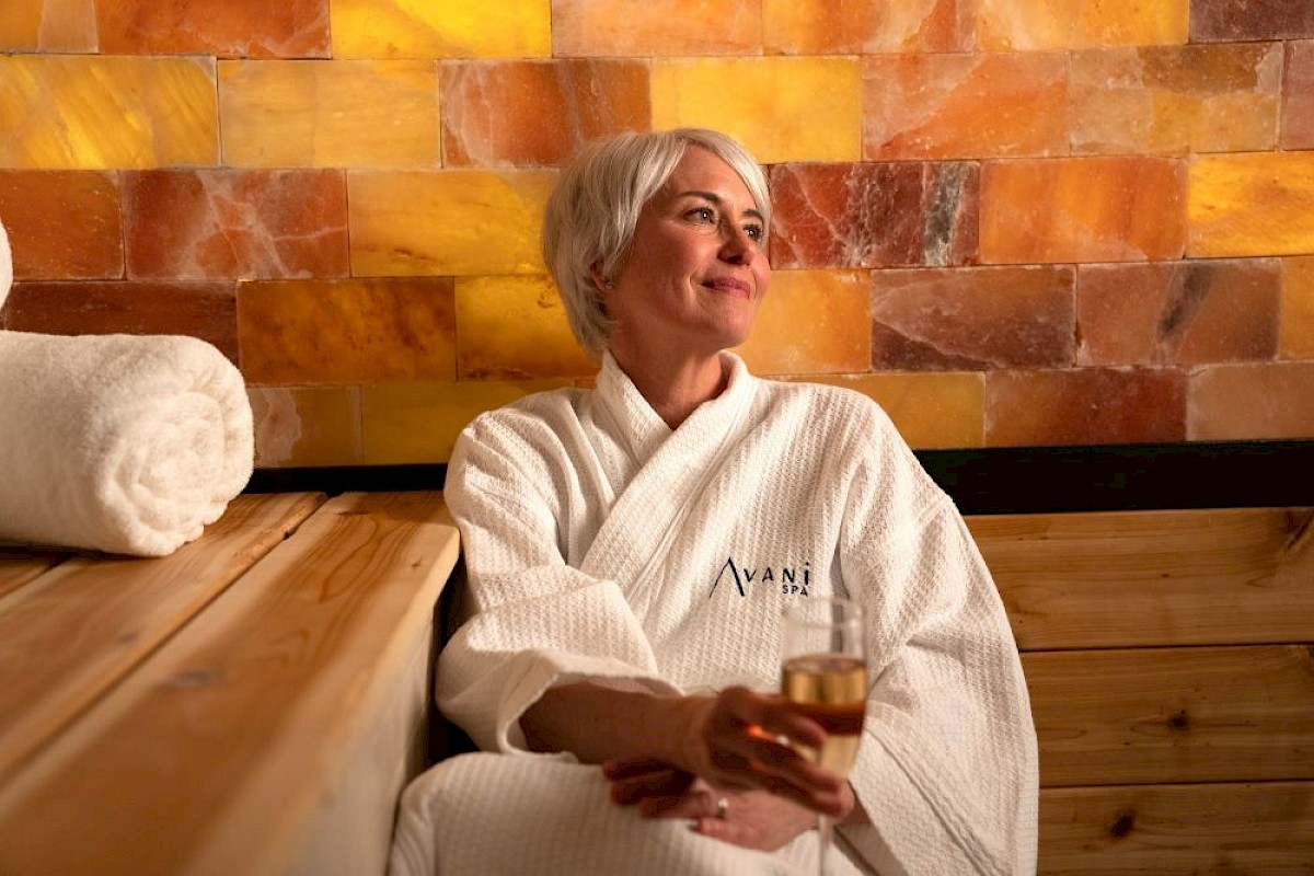 A person in a white robe relaxes with a drink against a warm, textured wall, next to rolled towels, enjoying a serene moment.