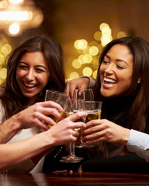 A group of five people is smiling and clinking their drinks together in a festive, warmly lit setting, likely celebrating or socializing.