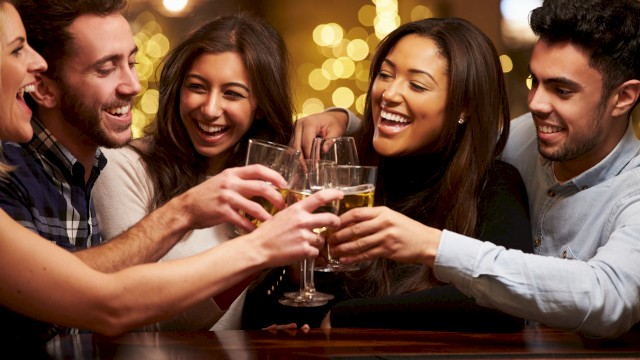A group of five people is smiling and clinking their drinks together in a festive, warmly lit setting, likely celebrating or socializing.