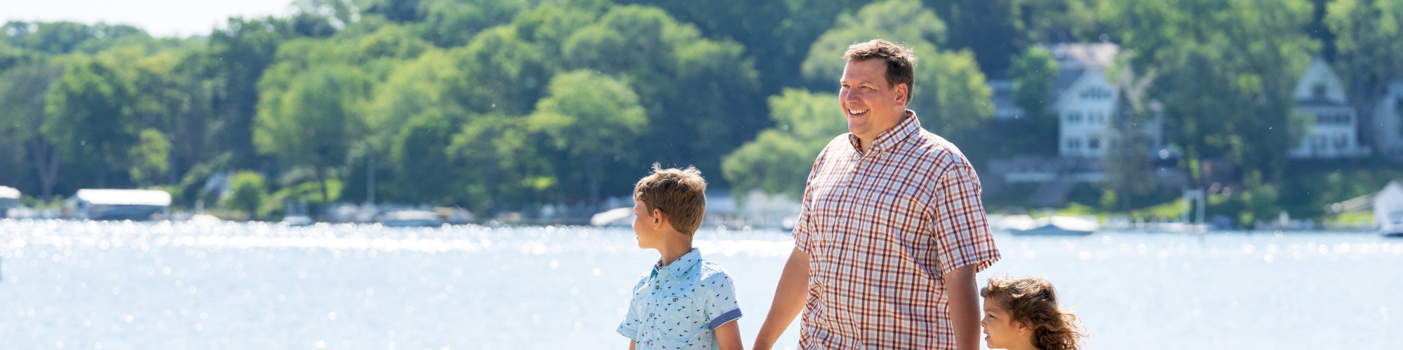 A man walks hand in hand with two children on a sunny beach path near a lake, surrounded by greenery and houses in the background.
