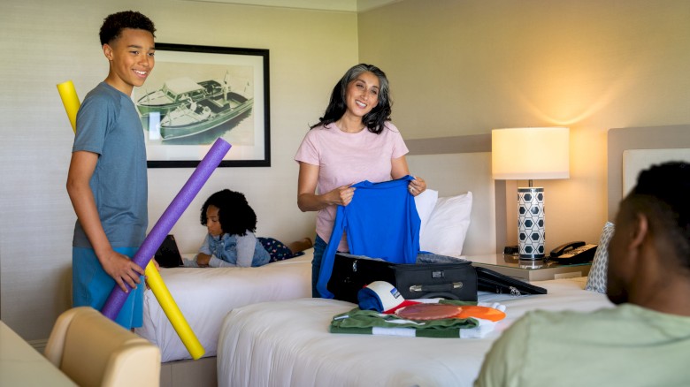 A family packing in a hotel room; a woman holds clothes, a boy has pool noodles, and a man and child are in the background.