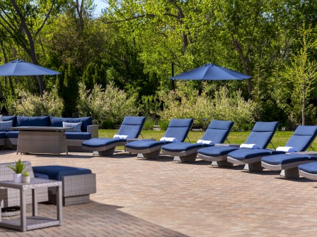 The image shows a patio area with blue lounge chairs, tables, umbrellas, and trees in the background.