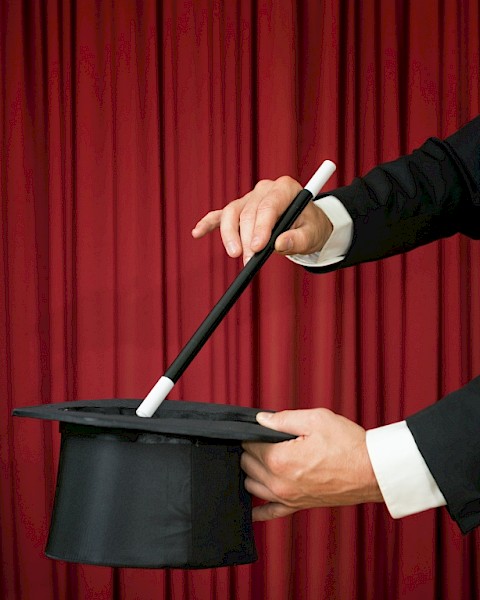 A magician is holding a top hat and a wand against a red curtain backdrop, suggesting a magic trick about to be performed.
