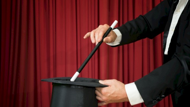 A magician is holding a top hat and a wand against a red curtain backdrop, suggesting a magic trick about to be performed.