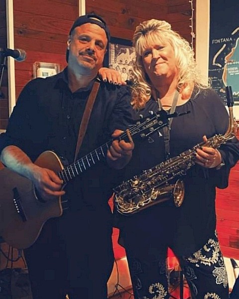 A man with a guitar and a woman with a saxophone pose together indoors, marked 