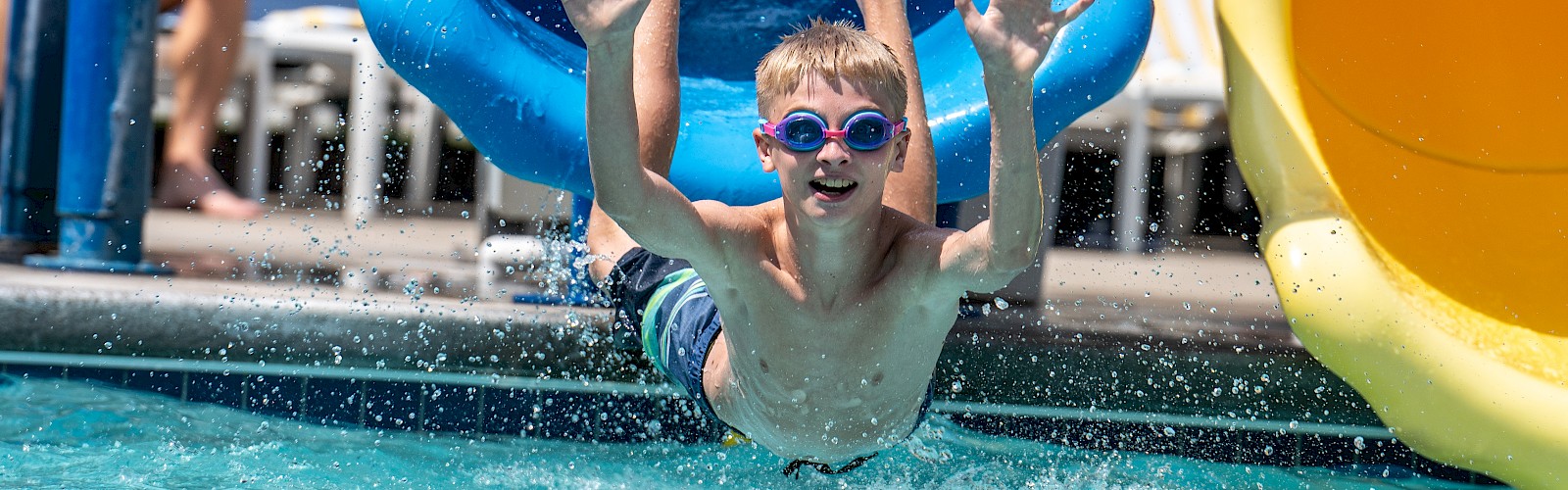 A child is joyfully coming out of a blue water slide into a pool.