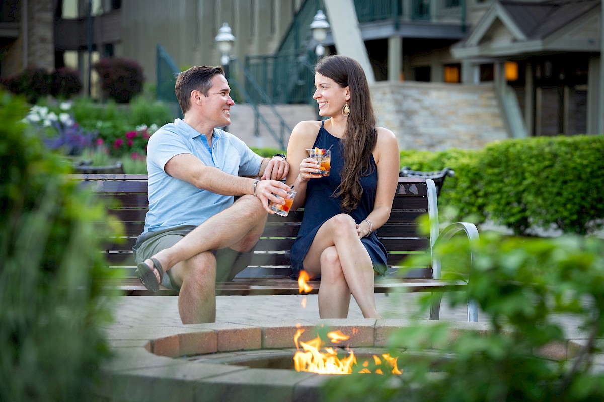 A man and woman sit on a bench by a firepit, enjoying drinks and each other's company in a lush outdoor setting.