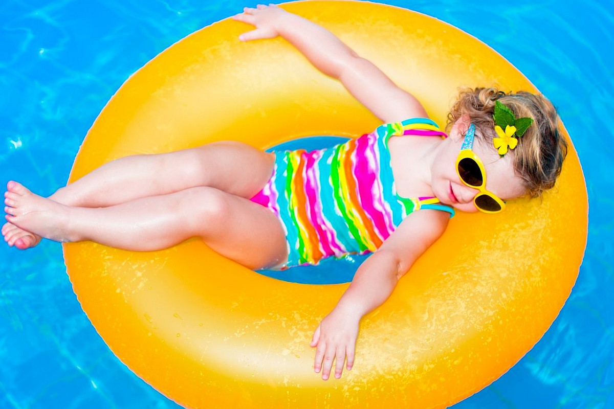 A young girl in sunglasses relaxes on a yellow float in a swimming pool, wearing a colorful striped swimsuit and a flower hair accessory.