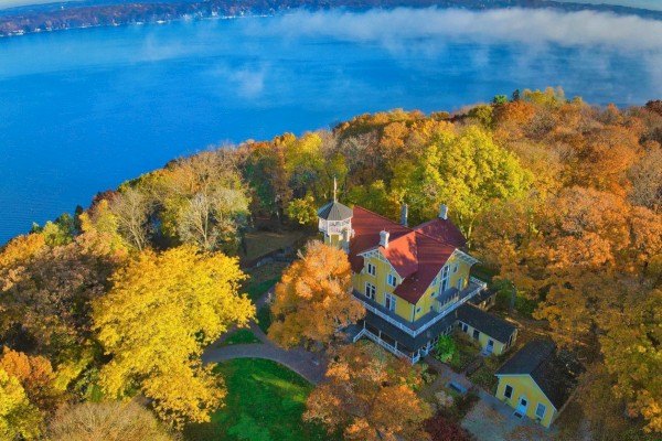 An aerial view of a yellow house surrounded by autumn trees near a large body of water under a clear, blue sky.