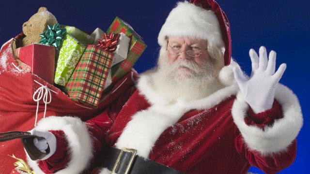 A person dressed as Santa Claus is waving, with a large red sack filled with wrapped gifts and a teddy bear over their shoulder, against a blue background.