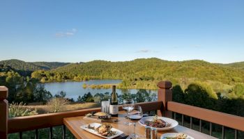 A wooden table with plates of food, a bottle of wine, and glasses overlooks a scenic lake surrounded by green hills under a clear blue sky.