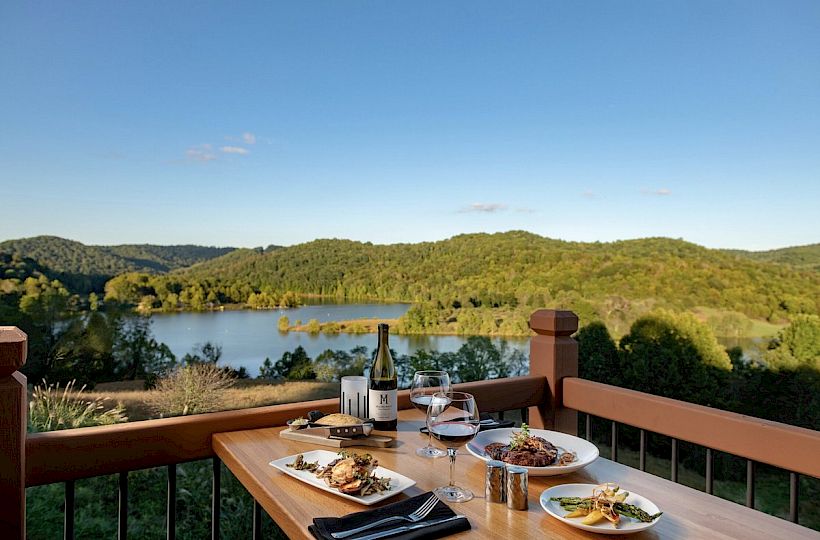 A wooden table with plates of food, a bottle of wine, and glasses overlooks a scenic lake surrounded by green hills under a clear blue sky.