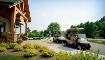 The image shows two golf carts near a building with wooden beams and stone walls. People are preparing for a golf game amid greenery.